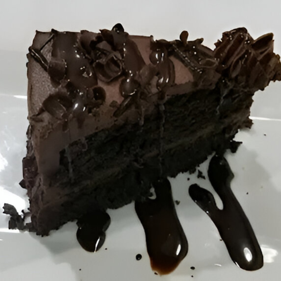 A rich slice of chocolate cake with chocolate frosting, shavings, and syrup on a white plate.
